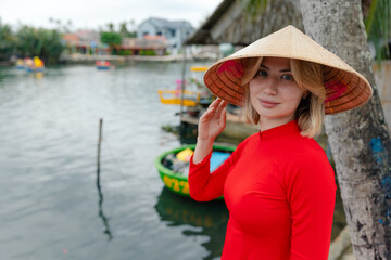 Fototapeta premium Young caucasian female tourist in ao dai at hoi an riverside, vietnam. Traveler in red traditional dress on coconut boat