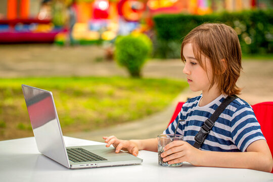 Teen boy with a laptop sits behind a table in a park. School boy is typing on computer outdoors. Child boy studying. Thoughtful schoolboy studying online on computer outdoors in park - Powered by Adobe