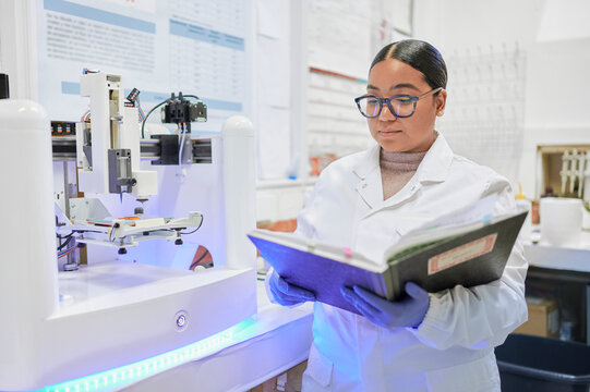 Latina woman scientist working in an academic lab checks her notebook beside a 3D printer while wearing a lab coat and glasses