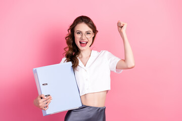 Naklejka premium Cheerful young woman holding a binder against a pink backdrop, celebrating success and radiating enthusiasm and style