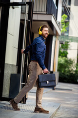 Fototapeta premium Handsome professional man exiting a modern office building, carrying a briefcase and wearing headphones in a vibrant cityscape