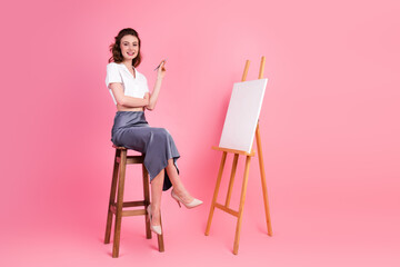Young elegant woman seated stylishly with an easel on pink background, embracing creativity and...