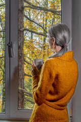 Woman with grey hair in cozy sweater gazes outside while holding mug, observing vibrant autumn...