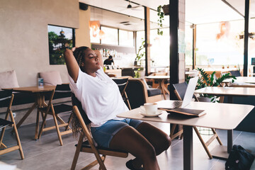 Black woman leans back eyes closed with arms behind head, enjoying moment of success and peace after completing digital freelance work in modern cafe.
