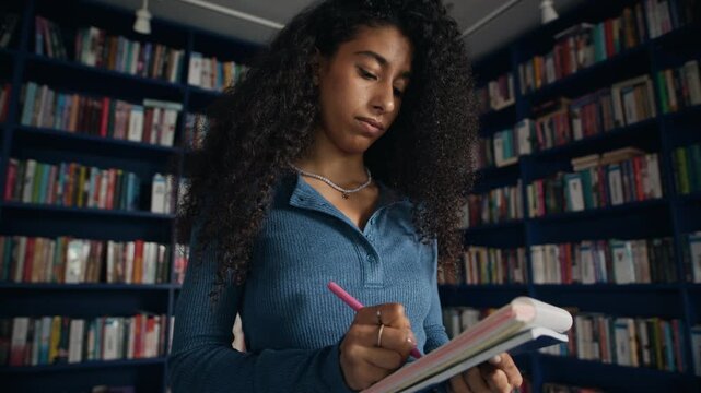 Medium shot of concentrated biracial female student standing in modern library in front of bookshelves and taking notes in her notebook, preparing list of sources for her research paper