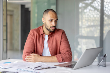 Focused young man wearing a casual shirt working remotely on his laptop in a modern environment with documents nearby. The setting suggests productivity, professionalism, and a relaxed workspace.
