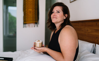Freelancer woman holding latte in bright room, laptop in view, expressing clarity, focus and...