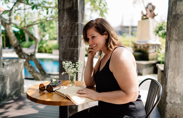 Freelancer woman smiling while looking up during creative journaling at tropical terrace, enjoying balanced moment of remote work and inner reflection.