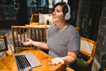 Smiling midlife freelancer enjoying online conversation with headphones, arms open in friendly...