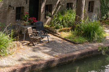 Outdoor seating area with two wooden chairs beside tranquil canal, surrounded by lush greenery and...