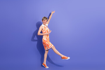 Young fashionable woman with short blonde hair in vibrant orange dress holding phone against purple...