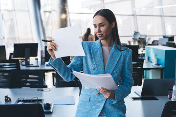 Businesswoman reviews printed documents while standing, expressing modern multitasking, flexible...
