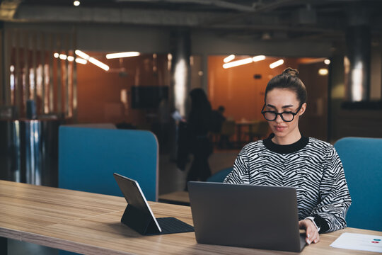 Businesswoman checks smartphone while multitasking with laptop and tablet in dim coworking space, illustrating modern mobile workflow and digital independence in creative office.