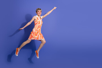 Obraz premium Energetic young woman jumps enthusiastically against a vivid purple background wearing a trendy orange dress and sneakers