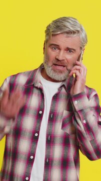 Mature man speaks on mobile phone with forceful tone, revealing anger agitation during tense talk quarrel. Middle-aged guy on yellow background shows argumentative stance, emphasizing heated dispute.
