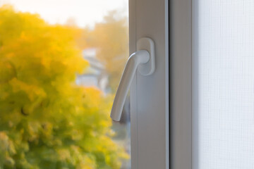 Window with a white handle and a white frame. The handle is open and the window is slightly ajar. The window is next to a tree with yellow leaves