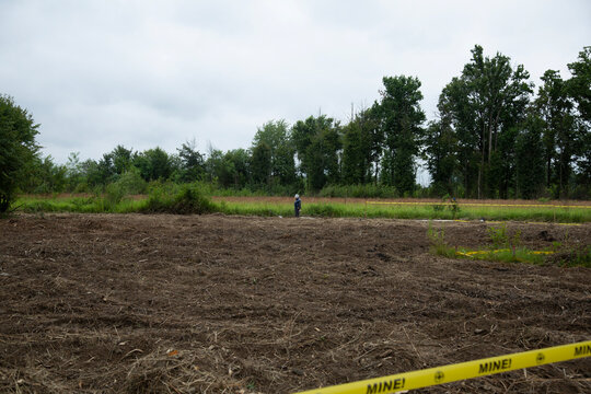 A documentary scene depicting a demining operation in a rural field marked with yellow mine warning tape, capturing the tension of explosive clearance.
