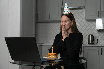 Young woman wearing party hat making a wish holding hands together looking at laptop celebrating...