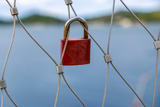 A red love lock with a heart symbol locked onto a steel cable fence, representing eternal love and commitment, with a blurred sea background.A romantic tradition - Powered by Adobe