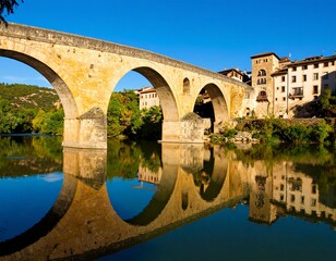 Fototapeta premium A charming scene of an old stone bridge over a river, with the reflection of the village houses perfectly captured in the water, creating a symmetrical view.