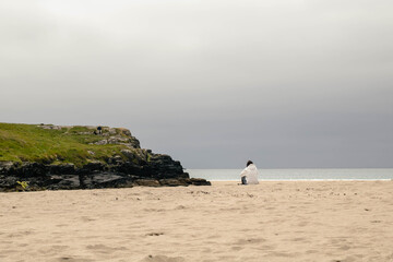 A lonely faceless woman in shapeless clothes sits thoughtfully on the beach, Barley Cove Beach,...