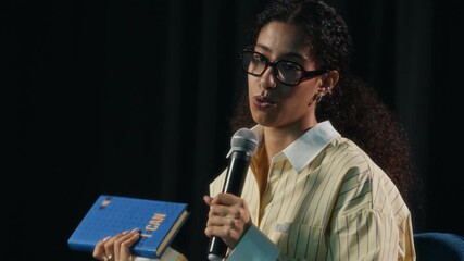 Tilt up shot of young confident biracial female author sitting on stage and speaking into microphone while presenting her new motivational book to unrecognizable audience