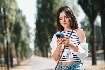 Young woman enjoying a sunny day at the park while texting on her phone, dressed in casual summer...
