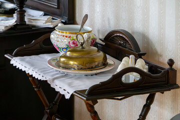 Antique wooden table with lace doily, brass cloche on plate, floral porcelain tureen, and salt shakers, retro-style interior.
