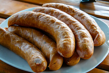 Traditional country sausages with golden smoked skin, arranged on a white plate on a wooden table, prepared for grilling, regional cuisine dish.
