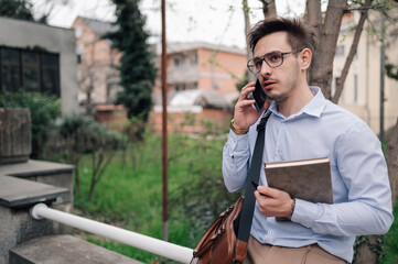 Businessman talking on phone while walking outdoors, holding notebook and pen
