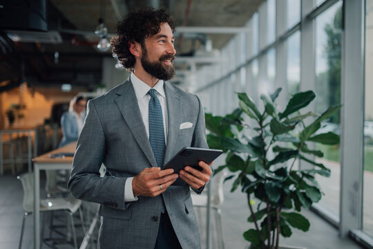 Smiling businessman holding tablet and looking away in modern office - Powered by Adobe