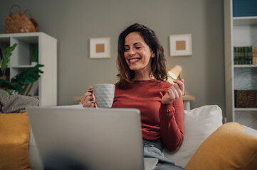 Happy woman drinking coffee and using laptop on sofa at home