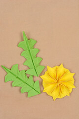 A yellow origami dandelion flower and its three green folded leaves are artistically arranged on a subtle brown background, highlighting the paper texture