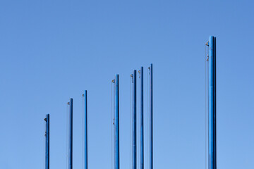 A series of empty blue flagpoles stand against a clear blue sky, creating a minimalist and striking...