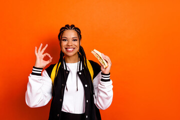 Fototapeta na wymiar Cheerful teenage student holding a sandwich and making an okay hand gesture against a bright orange background