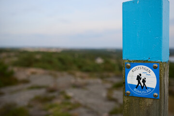 Close-Up of Kuststigen Hiking Trail Marker Near Str&ouml;mstad, Sweden with Coastal Landscape and Rocky Terrain in Soft Focus