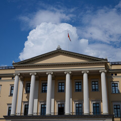 Neoclassical Facade of Oslo Royal Palace with Tall White Columns, Symmetrical Windows, and Norwegian Flag Under Blue Sky with Clouds