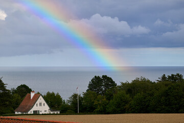 Colorful rainbow stretches above the Baltic Sea with a white house and trees in the foreground in Bornholm Denmark.
