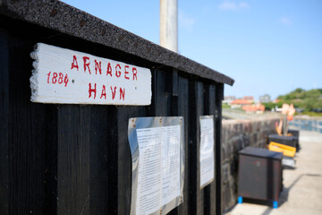 Close up of Arnager Havn harbor sign on wooden wall with informational boards and view of seaside pier on a sunny day in Bornholm Denmark