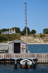 Small wooden boat moored at Arnager Havn pier with wooden shed, tall mast, and coastal landscape in Bornholm Denmark on a sunny summer day