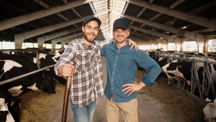 Two happy Caucasian male farmers working at farm standing in cowshed and smiling to camera. Joyful men work in barn together in village. Many cows in stable on background. Dairy industry. Livestock
