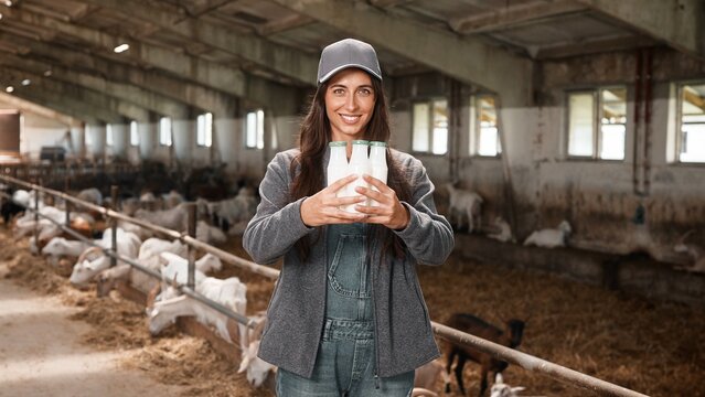 Portrait of cheerful young woman holding in hands bottles with milk standing inside the barn in village. Beautiful female farmer in animal farm carrying fresh ecological goat milk and smiling