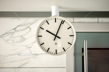 A minimalist white round clock with black hands hanging from a ceiling against a white marble wall....