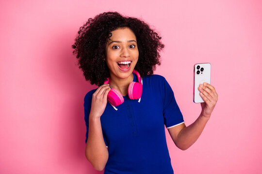 Excited young woman wearing pink headphones holding a smartphone against a pink background with a joyful expression