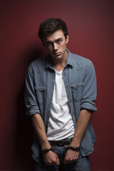 A young man faces the camera, handcuffed, wearing casual clothes against a red background, expressing seriousness.