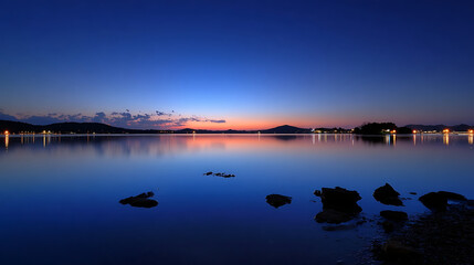 Fototapeta premium Tranquil lake at twilight with distant lights reflecting on calm water and dark rocks in foreground