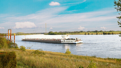 Fototapeta premium Tanker barge sailing on the Rhine River with a cable-stayed bridge in the background, transporting cargo on a sunny day in a scenic riverside landscape.