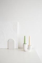Two candles in curved geometric candle holder and painting with white putty stand on chest of...
