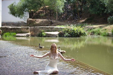 A young, beautiful blonde woman wearing a beige knitted dress sits by a small waterfall on a hot...