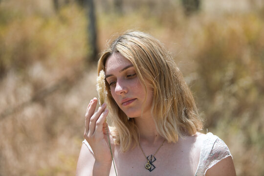 Portrait of a young, beautiful blonde woman with a serene gaze holding a dry, soft ear of corn in her hands and placing it next to her face with delicacy and tenderness.